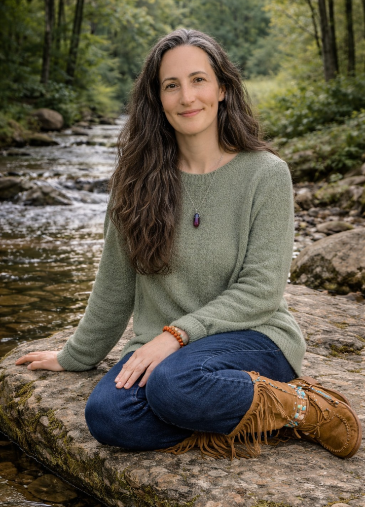 Woman wearing tall Native American boots, blue jeans and a sage green sweater who is sitting on a rock near a creek.